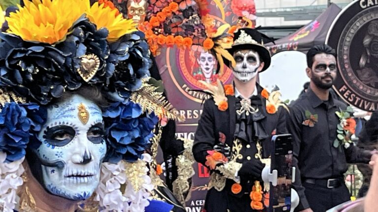 Festival scene showing Día de los Muertos skull makeup, large blue and yellow flower crown and decorated black attire